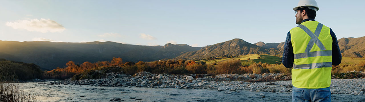 Técnico con casco y chaleco reflectante observa un río rodeado de montañas
